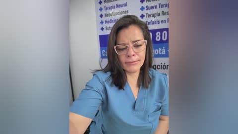 Media: Video of a middle-aged woman with light skin, shoulder-length dark hair, and glasses, wearing a blue scrub top, standing against a blue and white informational poster in a medical setting.