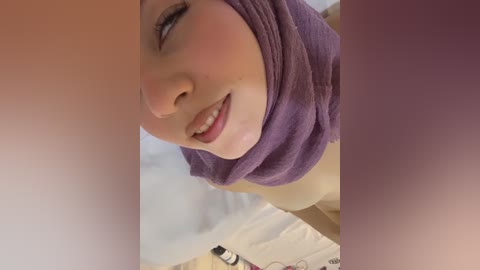 Video of a smiling Caucasian woman with light skin and blue eyes, wearing a purple hijab and a white shirt. Background shows a white bed with a black object on it.