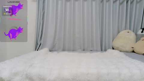 Video of a plush, white-furred cat laying on a fluffy, white rug in a room with light gray curtains. The room features a digital display with cat icons and \"Lovingly\" text in the background.