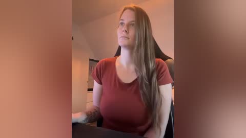 A video of a young Caucasian woman with long, straight brown hair, wearing a red short-sleeved top, sitting in an office chair. The background is a dimly lit room with neutral tones.