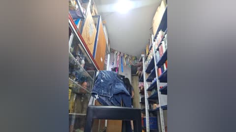 A video of a narrow, cluttered store with a glass display case on the left and shelves of colorful products on the right. A person in jeans and a jacket is bending over, likely organizing items.