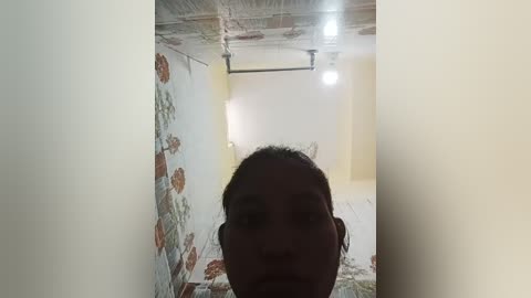 Video of a woman with dark hair and medium skin tone standing in a narrow, unfinished room with exposed white and gray ceiling, light fixture, and floral-patterned wallpaper.