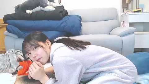 Video of an Asian woman with straight black hair, wearing a gray robe, lying on a blue sofa in a messy living room with stuffed toys and clothes scattered around.
