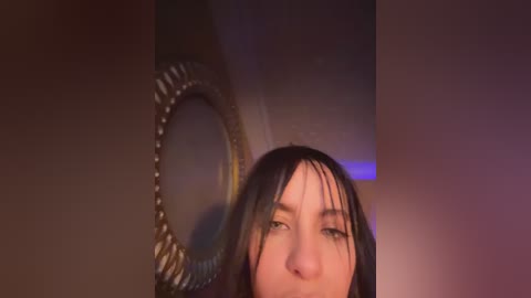 Video of a young woman with wet hair, partially obscured by a dimly lit, round, ornate mirror on a brown wall.