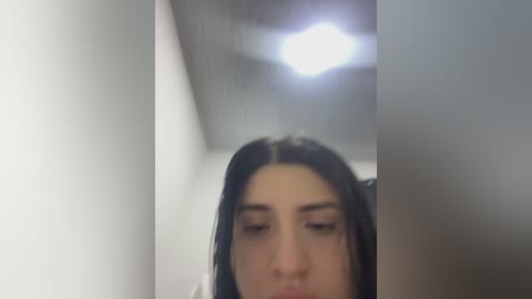 A blurred video of a young woman with long, dark hair and fair skin, captured in a dimly lit, narrow space with a bright light above.