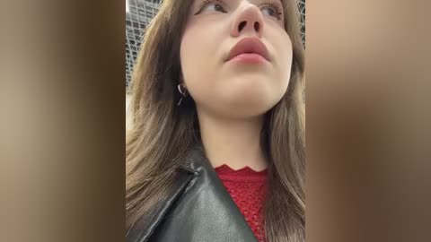 Video of a young woman with fair skin, brown hair, and light eyes, wearing a black leather jacket over a red lace top. Background features a blurred, mosaic-patterned wall.