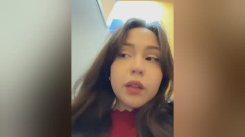 A close-up video of a young woman with long brown hair, fair skin, and red lipstick, wearing a dark jacket over a red top. She gazes forward with a neutral expression. The background is blurred, showing a dimly lit room with a ceiling light.
