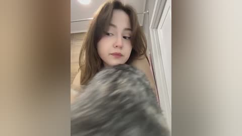 A video captures a young woman with light skin and shoulder-length brown hair, looking down at a fluffy grey cat. The background features a white door and beige tiles. The image is slightly blurred.