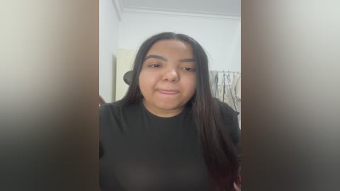 Video of a young woman with long dark hair, wearing a black top, standing in a dimly lit room with white walls and a beige curtain.
