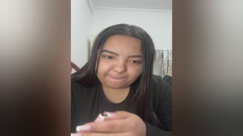 Video of a young woman with long black hair and light brown skin, wearing a black shirt, smiling while holding a smartphone in her hand. Background includes a white wall and a blurred hanging object.