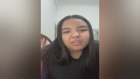 Video of a young woman with medium brown skin, straight black hair, and full lips, wearing a black shirt. She has a neutral expression, standing in a white-walled room with a wooden chair behind her.