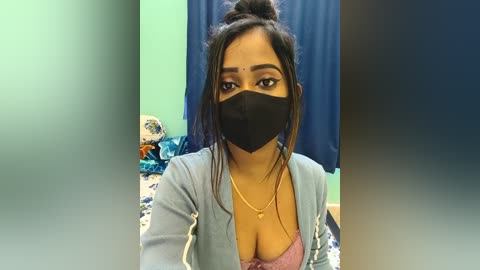 Video of a South Asian woman with medium skin tone, dark hair in a messy bun, wearing a black face mask, light blue cardigan, and a revealing purple bra, standing in a room with blue curtains and a bed.