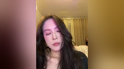 Video of a young Asian woman with long, dark hair and light skin, wearing makeup and a black top, smiling and looking down. Background features beige curtains and a bed.