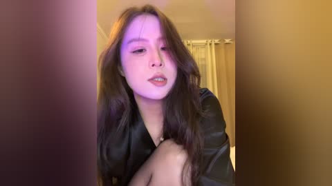 Video of a young woman with long, dark hair, wearing a black leather jacket, leaning against a beige wall in a dimly lit room.