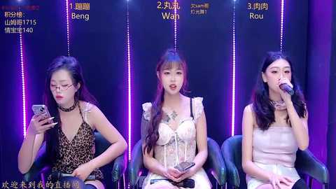 Video of three East Asian women, each seated in a black chair, singing into microphones. They wear different colorful dresses against a dark purple backdrop.