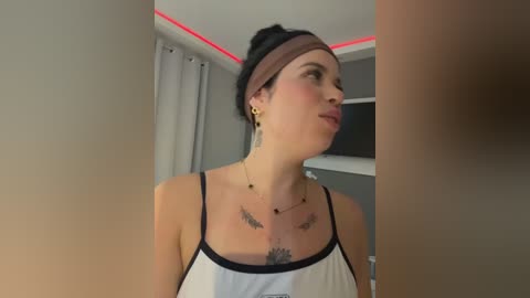 Video of a fair-skinned woman with dark hair in a bun, wearing a white spaghetti-strap top, gold earrings, and a headband, standing in a modern room with gray walls and red accent lighting.