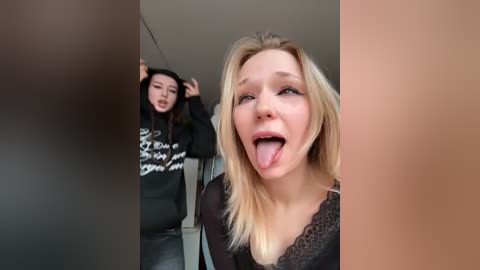 Video of a blonde woman with light skin, sticking out her tongue, in a black lace top, while an Asian woman in a black hoodie with white text stands in the background.