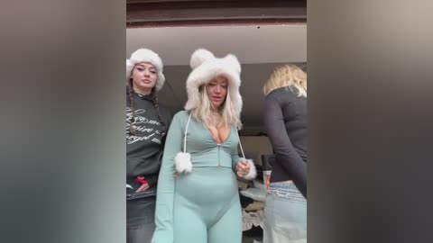 Video of two women in matching fluffy white cat ears headbands and light blue onesies. One woman, with fair skin and long blonde hair, has a large cleavage. The background shows a blurred, indoor setting with another woman in the distance.