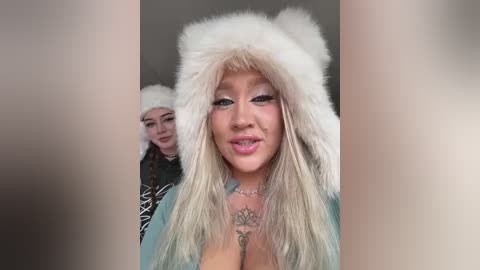Video of a blonde woman with a large bust wearing a white fur hat and a light blue shirt, standing in front of a blurred background with another person in the background.
