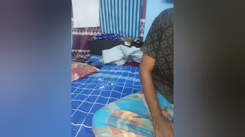 Video of a small, cluttered room with a person in a dark shirt kneeling on a blue-checkered bed, surrounded by colorful, patterned pillows and blankets.