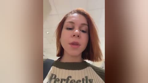 Video of a young woman with fair skin and straight, shoulder-length auburn hair. She wears a green and beige striped sweater with \"Perfectly\" printed on it. Background shows a white brick wall.