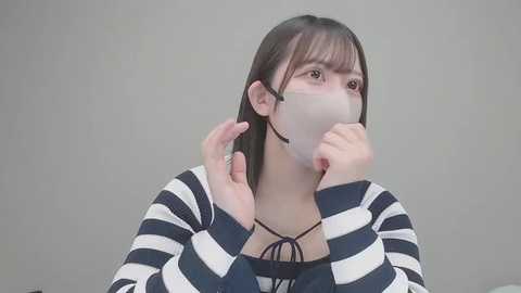 Video of an Asian woman with long, straight black hair, wearing a black-and-white striped shirt, white mask, and a black necklace, adjusting the mask with both hands, against a plain gray background.