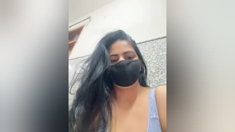 A video of a young woman with long black hair and light brown skin, wearing a black face mask and a light blue tank top, taken indoors with a blurred background.