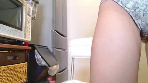 Video of a woman's bare right thigh and lower leg, wearing floral-patterned shorts, standing in a cluttered kitchen with a microwave, oven, and a white chair.