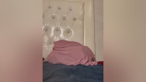 Video of a neatly made bed with a plush, tufted white headboard adorned with silver buttons. A soft pink blanket covers the bed, resting on a dark blue bedspread. The room's walls are plain and beige.