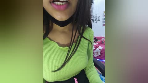 Video of a young woman with medium brown skin and straight black hair, wearing a neon green ribbed crop top and black choker, smiling. Background shows a red floral bedspread.