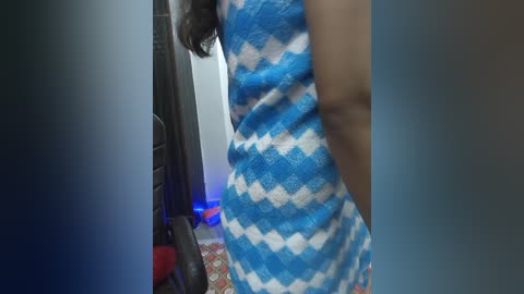 Media: A video of a woman in a blue and white diamond-patterned dress, standing in a dimly lit room with a red-patterned rug and a black chair.