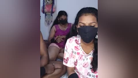 Video of three South Asian women, wearing masks and pink clothes, sitting on a bed in a dimly lit room with a stuffed animal on the wall.