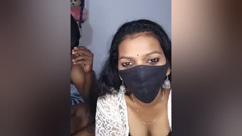 Video of a South Asian woman with medium-dark skin, long black hair, and wearing a black face mask, white lace top, and dangling earrings, holding hands with another person.