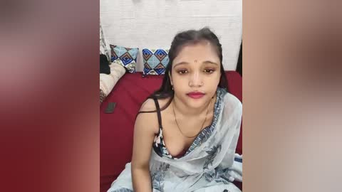Video of a South Asian woman with medium brown skin, wearing a semi-transparent white sari, seated on a red bed, with patterned pillows and a white brick wall background.