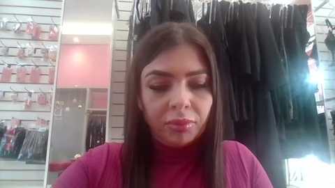 Video of a young woman with long, straight brown hair, wearing a pink turtleneck sweater, applying makeup. Background features a mirror, shelves with cosmetics, and a black dress on a hanger.