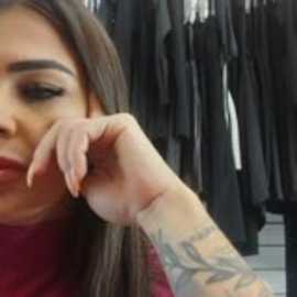 Video of a Latina woman with long brown hair, dark eyes, and red lipstick, touching her face. She wears a red top and has colorful tattoos on her arm. Background shows hanging black clothes.