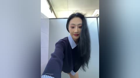 A video of an Asian woman with long black hair, red lipstick, and wearing a navy sweater with a white collar, leaning against a frosted glass wall in a modern office.