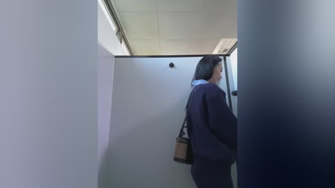 A video of an Asian woman with long black hair, wearing a dark blue suit and carrying a brown handbag, walking through a narrow, dimly-lit hallway with white walls and fluorescent lights.