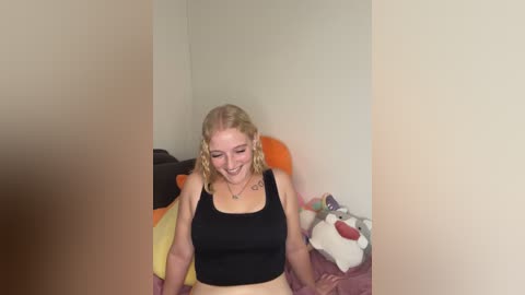 Video of a smiling blonde woman in a black crop top, with a tattoo on her left arm, in a cluttered bedroom with stuffed animals.