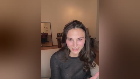 Video of a young woman with long brown hair, wearing a black top, seated on a beige couch in a cozy, warmly lit room with a mirror and wooden furniture in the background.