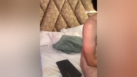 Video of a person sitting on a bed with a tufted, patterned headboard, wearing a teal robe and black keyboard on white sheets.