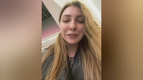 Media: Video of a young woman with long, blonde hair, wearing a black leather jacket, smiling slightly, taken indoors with a blurred, beige wall and ceiling in the background.