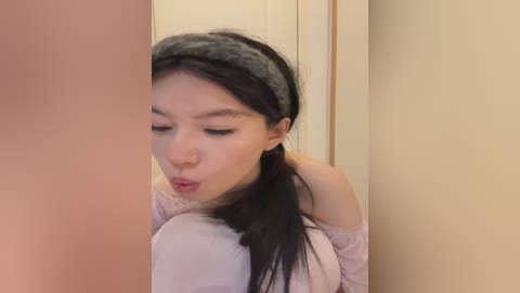 Video of a young Asian woman with long black hair and fair skin, wearing a light pink off-shoulder top, blowing a kiss in front of a beige wall.
