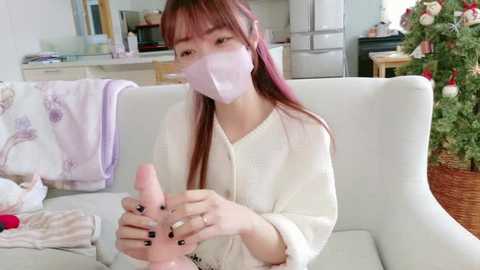 A video of an Asian woman with long, straight brown hair, wearing a white robe and pink face mask, massaging a large, erect penis in her hands in a modern living room.