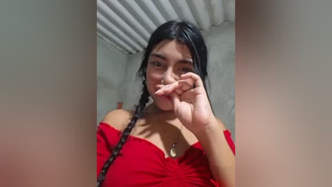 A video of an Asian woman with long black braids, wearing a red off-shoulder dress, covering her mouth with her fingers, standing in a room with a textured ceiling.