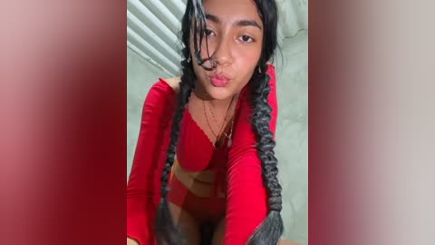 Video of a young woman with medium brown skin, wearing a red long-sleeve top and matching bralette, with long black braids, pouting her lips. Background shows white blinds and a light-colored wall.