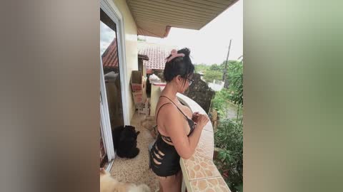 Video of a woman in a black, ripped swimsuit on a balcony, adjusting her hair. The background features a beige wall and a green shrub.