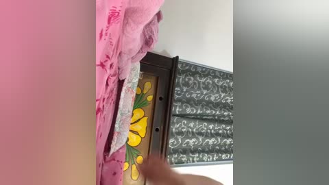 A video of a person's hand delicately peeling off a pink fabric with intricate floral patterns, revealing a decorative mirror with floral motifs and a silver, ornate door frame in the background.