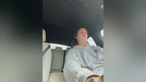 Video of a woman with shoulder-length brown hair, wearing a light gray hoodie, sitting in the backseat of a car with beige upholstery and tinted windows.