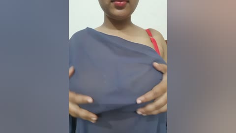 Video of a person with medium-dark skin, wearing a red bra and a sheer blue top, holding their breasts. The background is blurred and white.